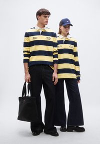 Ριγέ πόλο μπλούζες σε ναυτικό μπλε και κίτρινο, σκούρο τζιν παντελόνι και μαύρη τσάντα tote. Διαθέτει γιακά σε αντίθεση και κεντημένο λογότυπο.