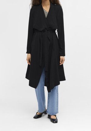 Woman wearing a long black belted coat over light blue jeans and black low-heeled shoes, standing against a plain background.