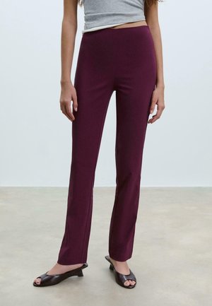 Femme portant un pantalon violet ajusté et des chaussures noires à bout ouvert à talon bas, debout sur un sol uni avec un fond gris clair.