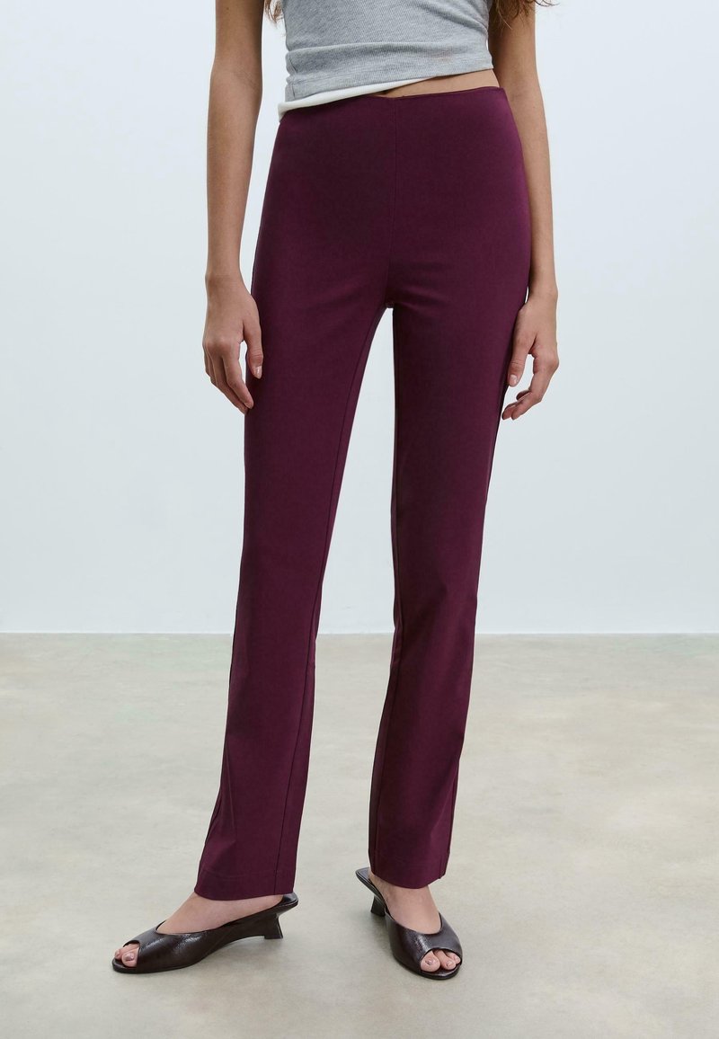 Femme portant un pantalon violet ajusté et des chaussures noires à bout ouvert à talon bas, debout sur un sol uni avec un fond gris clair.