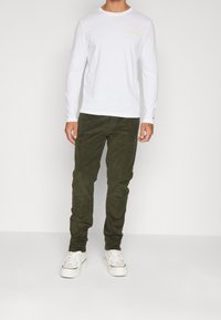Vit bomullströja med långa ärmar och gultexten "HILFIGER" på bröstet, matchad med olivgrön kordflorsebyxor och vita sneakers.