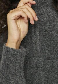 Šedý vlněný svetr s vysokým límcem a texturovaným povrchem. Ruka drží zip, aby zdůraznila měkkost a detaily látky.