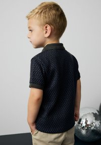 Polo shirt a righe navy con colletto verde scuro, maniche corte e tessuto testurizzato. Mano di un bambino infilata nella tasca laterale di pantaloni beige.