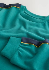 Zöldeskék pulóver, sötétkék és sárga csíkos díszítéssel a vállán és bordázott mandzsettával világos háttéren.