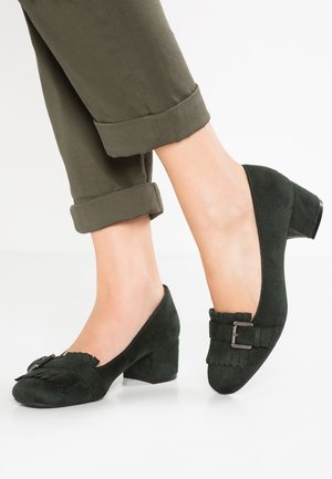 Buty z ciemnozielonego zamszu z zaokrąglonym noskiem, falistymi krawędziami i ozdobną klamrą. Gruby obcas dodaje wysokości i stabilności.