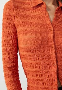 Camicetta button-up arancione rustico con un motivo ondulato testurizzato, dotata di maniche lunghe e colletto. I bottoni sono dello stesso colore del tessuto.