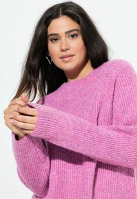 Rosa stickad tröja med ribbat mönster, nedhasade axlar och långa ärmar. Har en rund halsringning och en avslappnad passform.