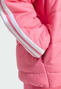 Close-up of a cotton candy pink puffer jacket, featuring white stripes along the sleeve, with a hand resting in the pocket.