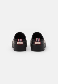 Juodos gumos slip-on tipo batai su pakeltu padu, turintys raudoną, baltą ir juodą dryžuotą traukimo juostelę bei raudoną "HUNTER" logotipą gale.