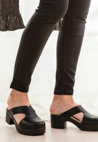 Zwarte leren muiltjes met een open achterkant en uitsneden. Dikke hak en platformzool bieden ondersteuning. Gecombineerd met zwarte skinny jeans.