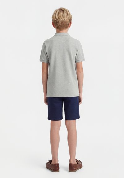 Garçon blond debout de dos, portant un polo gris clair à manches courtes, un short bleu marine et des chaussures marron sur fond blanc.