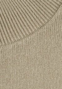 Close-up van beige gebreide stof met twee duidelijke geribbelde patronen die elkaar kruisen bij een diagonale naad.