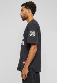 Tricou de baseball negru cu mâneci scurte, având inscripții bej și un logo circular pe mâna stângă. Material texturat și croială lejeră.
