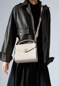 Sac crème en cuir embossé crocodile avec des accessoires dorés, de forme structurée, poignées supérieures et bandoulière amovible, porté avec une veste en cuir noir.