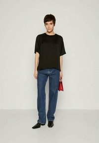 Young person standing wearing black short-sleeve top, blue jeans, black pointed shoes, holding small red handbag by side against plain background.