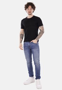 Camiseta negra de manga corta, jeans ajustados de color azul claro y zapatillas blancas. El modelo muestra tatuajes en los brazos, en una pose de pie con las manos en los bolsillos.