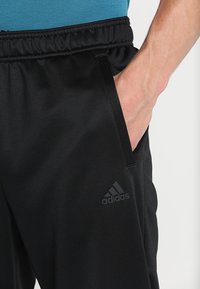 Personne portant un pantalon Adidas noir avec une taille élastique, main droite dans la poche latérale, portant une chemise bleue.