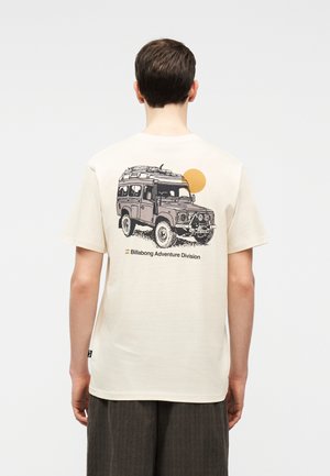 Osoba ubrana w kremową koszulkę z ilustracją pojazdu terenowego, żółtym słońcem i napisem "Billabong Adventure Division" na plecach.
