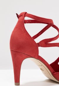 Sandale à talons hauts en daim rouge avec des brides croisées, bout pointu et un talon fin et effilé. L'intérieur présente un branding subtil.