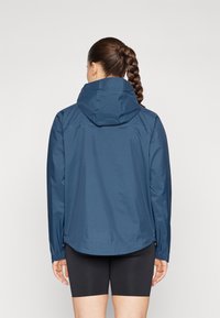 Marineblauwe waterdichte jas met een capuchon, voorzien van een gestructureerd oppervlak, lange mouwen en verstelbare zijzomen. Zwarte shorts worden eronder gedragen.