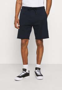 Nachtblaues Baumwollshorts mit elastischem Taillenbund und Kordelzug. Mit abgerundetem Saum und Seitentaschen. Kombiniert mit schwarzen Sneakers und weißen Socken.