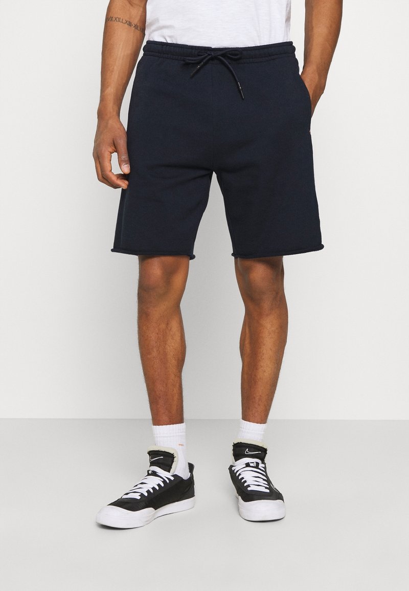 Nachtblaues Baumwollshorts mit elastischem Taillenbund und Kordelzug. Mit abgerundetem Saum und Seitentaschen. Kombiniert mit schwarzen Sneakers und weißen Socken.