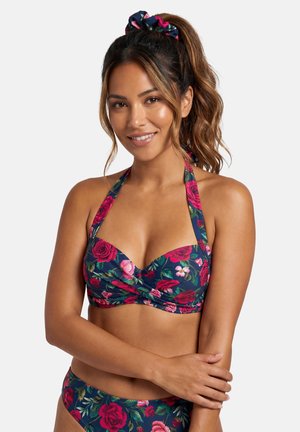 Kobieta o długich włosach, ubrana w granatowy bikini w kwiaty z wiązaniem na szyi i dopasowane majtki, uśmiechająca się z założonymi na piersi ramionami.