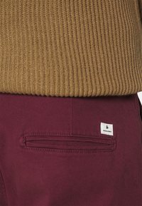 Jersey marrón con canalé superpuesto a pantalones marrones con un bolsillo trasero visible y una pequeña etiqueta blanca con el nombre de la marca "Jack & Jones."