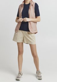 Lätt rosa ärmlös väst över en mörkblå ribbad skjorta, i kombination med beige shorts och gråa träningsskor med vita detaljer.