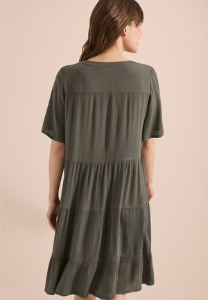 Kvinde med brunt hår iført en løs, lagdelt, struktureret olivengrøn kjole med korte ærmer, vist bagfra mod en beige baggrund.