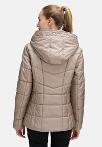 Beżowa pikowana kurtka z kapturem, pikowana faktura i pionowe szwy. Posiada dopasowaną sylwetkę i wysoki kołnierz. Widoczne czarne spodnie.