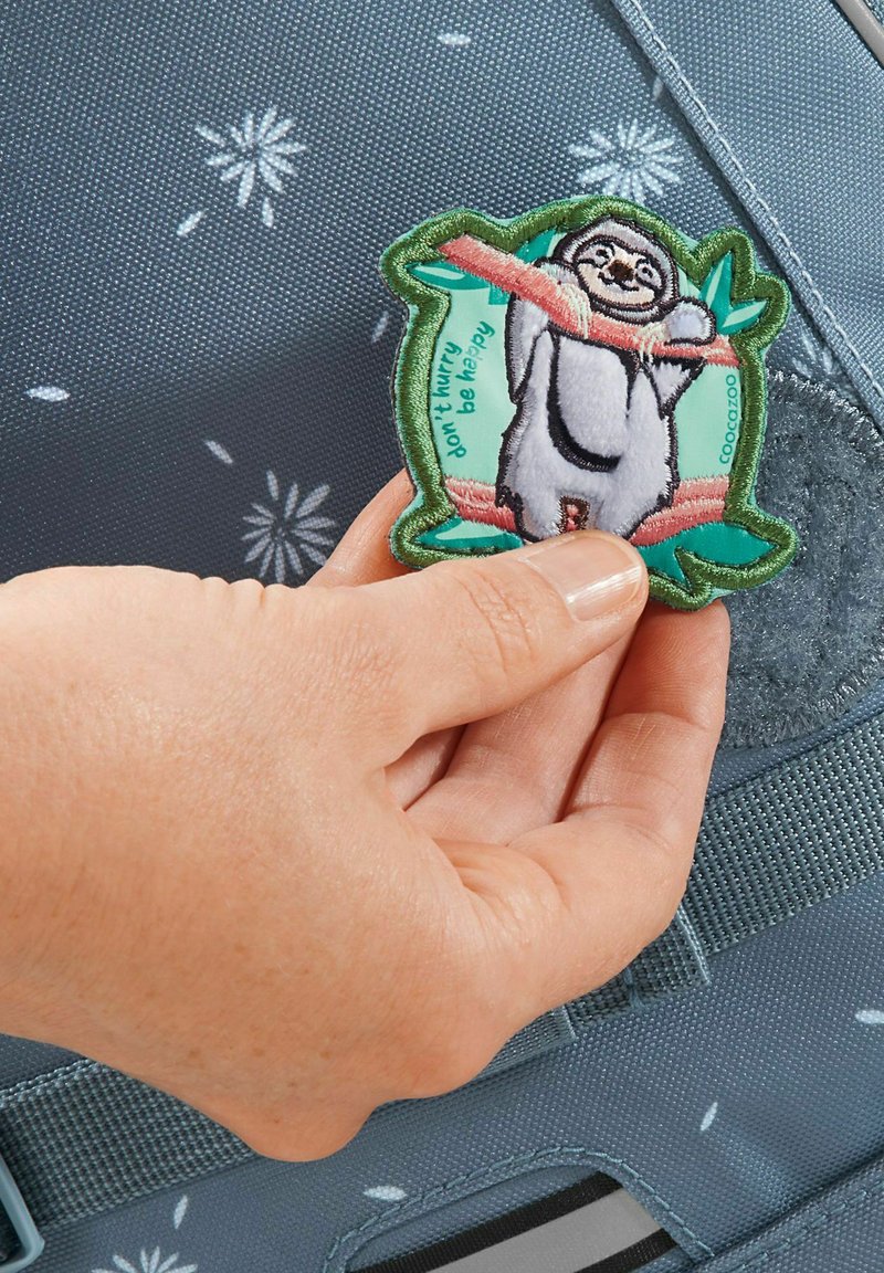 Un patch brodé coloré représentant un paresseux suspendu à une branche, sur un fond bleu texturé avec des motifs floraux blancs.