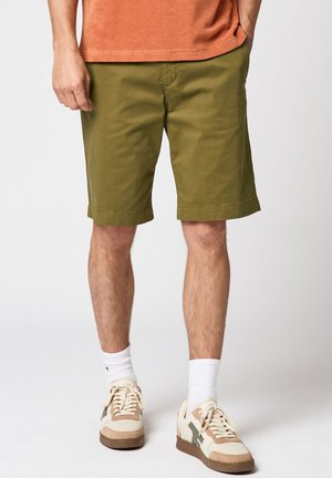 Mann trägt olivgrüne Shorts, weiße Sportsocken, beige-braune Sneaker und ein orangefarbenes Hemd, steht mit den Händen in den Taschen.