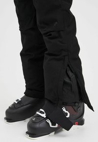 Black ski pants made of durable fabric, featuring zippered side openings. Paired with black ski boots that have a white buckle and patterned accents.