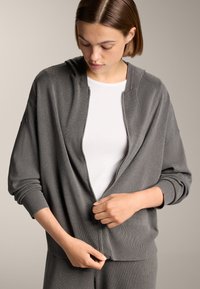 Grauer Reißverschluss-Hoodie mit lockerer Passform, gerippten Bündchen und Saum sowie einer weichen Textur. Getragen über einem weißen Oberteil, mit Kapuze und offener Vorderseite.