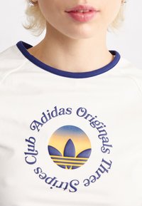 Bílý tričko s modrým výstřihem, s kulatým logem v gradientních barvách a nápisy "Adidas Originals" a "Club Sneakers" kolem něj.