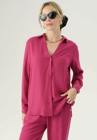 Camicia fucsia con bottoni e colletto, maniche lunghe, realizzata in tessuto liscio. Presenta un collo a V e bottoni rosa, indossata con pantaloni abbinati.