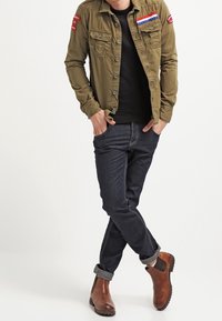 Olivgrüne Button-Up-Hemd mit Brusttaschen und Aufnähern, schwarzer Pullover darunter, dunkle Jeans, braune Stiefeletten mit elastischen Seiten.