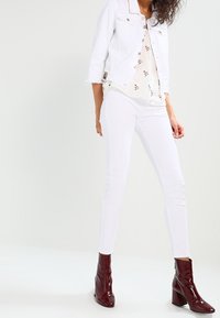 Chaqueta de mezclilla blanca con puños deshilachados sobre una blusa de estampado floral. Pantalones blancos ajustados combinados con botas de tobillo burdeos.