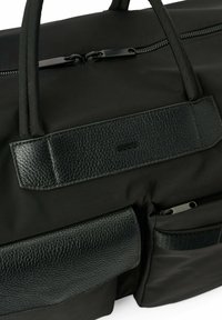 Musta duffel-laukku, joka on valmistettu kestävästä kankaasta ja jossa on teksturoitu nahkaakkentti, varustettu vetoketjulla ja sivutaskuilla säilytystä varten.