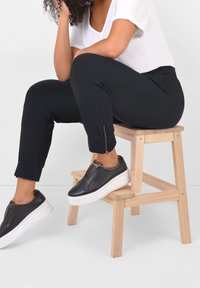 Svarta joggerbyxor med dragkedjeslut, bärs med svarta slip-on skor med vit sula. Toppdelen är en avslappnad vit t-shirt.