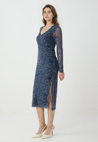 Abito blu navy con maniche lunghe trasparenti, motivo a pois multicolore, scollatura a V e spacco laterale. Abbinato a scarpe con tacco nudo.