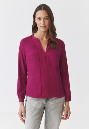 Vibrant magenta blouse with a v-neckline, long sleeves, and subtle pleats. Soft fabric with a smooth texture, paired with light gray jeans.