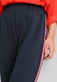 Námořnické kalhoty vysokého pasu s elastickým pasem, se červenými a bílými bočními pruhy a hladkou, lehkou texturou látky.