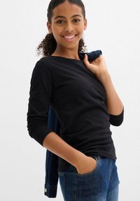Maglietta nera a maniche lunghe, in tessuto morbido, design aderente, indossata con jeans in denim blu, con una tasca, modello sorridente, che tiene una giacca.