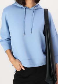 Sudadera con capucha de color azul claro y corte relajado, con cordones y una bolsa negra texturizada descansando en el hombro.