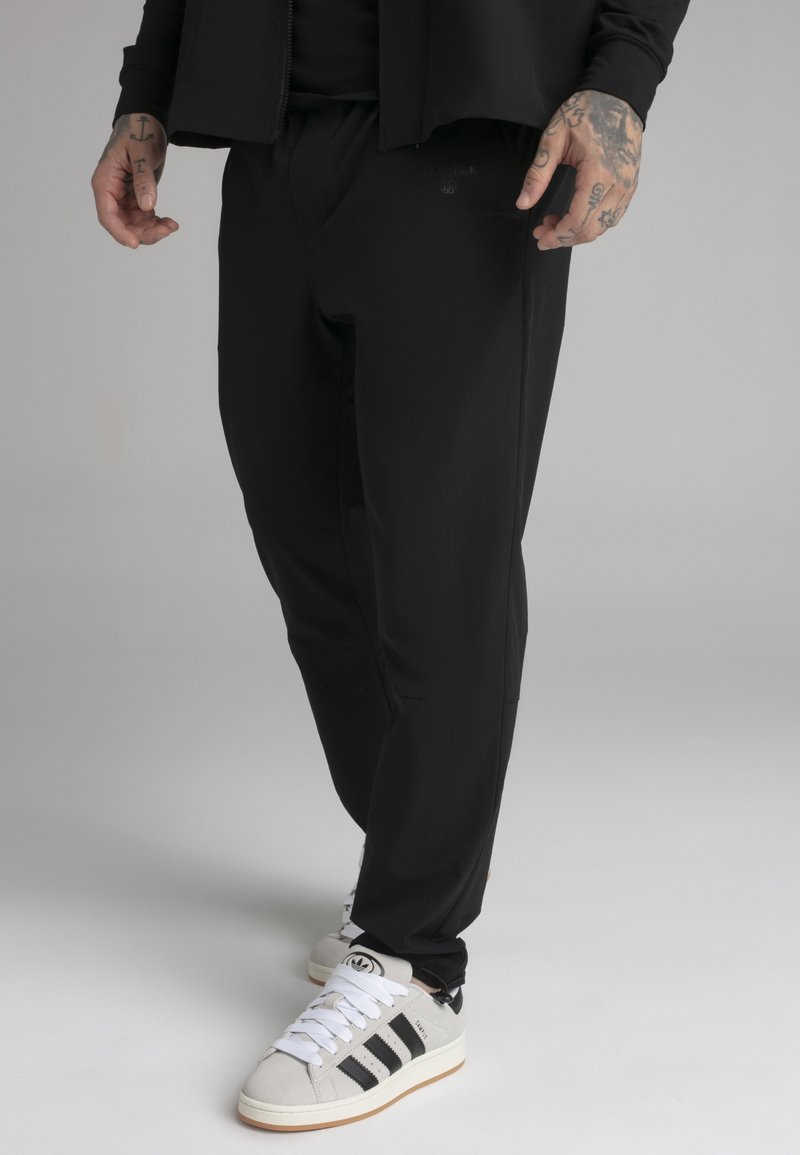 Homme portant un pantalon noir et des baskets blanches Adidas avec des bandes noires, debout sur un fond gris, montrant ses mains et poignets tatoués.