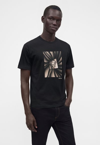 Άνδρας που φοράει μαύρο t-shirt Versace με κεντρικό σχέδιο κεφαλής Μέδουσας και σπειροειδές φόντο, στέκεται με το ένα χέρι στην τσέπη μπροστά σε απλό φόντο.