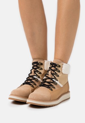 Bottes d'hiver en cuir tan à lacets pour femmes, avec doublure en fausse fourrure blanche, portées sur des jambes nues contre un fond blanc.