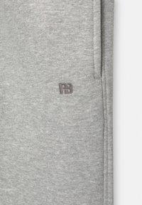 Tissu gris clair avec une couture verticale et un petit logo brodé près de la couture en fil gris plus foncé.